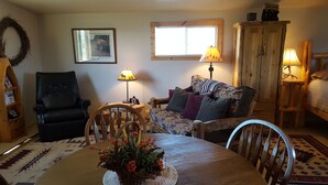 Un salon chaleureux avec une table à manger en bois, un fauteuil en cuir noir, un canapé à motifs et un buffet en bois.