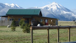 Une cabane en bois avec un toit vert, entourée d’une clôture en bois, se dressant sur un fond de montagnes enneigées.