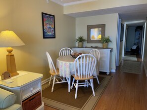 A dining area with a table set for four, white chairs, a lamp, a framed poster, and a dresser with potted plants. - The Leeway, Beautiful Gulf Front Condo on the Gulf of Mexico (Gulf Shores)