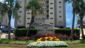 A high-rise building named "The Gulf Tower" with multiple balconies, surrounded by palm trees and a landscaped garden with various flowers. - The Leeway,  Beautiful Gulf Front Condo on the Gulf of Mexico (Gulf Shores)