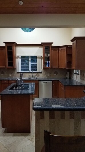 A kitchen with wooden cabinets, a granite countertop, a built-in dishwasher, and a window with blinds.
