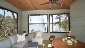 A room with a wooden ceiling, large windows, a grey sofa, a wicker chair, a wooden dining table with glasses, and a view of a lake.