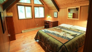 A bedroom with a wooden ceiling, a large bed with a quilt, a nightstand with a lamp, and a window with a view of trees. - CABIN LOOKING ACROSS THE SKY RIVER AT THE WILD SKY WILDERNESS (Skykomish)