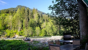 An outdoor deck with a grill and seating area overlooking a river and forested mountains. - CABIN LOOKING ACROSS THE SKY RIVER AT THE WILD SKY WILDERNESS (Skykomish)