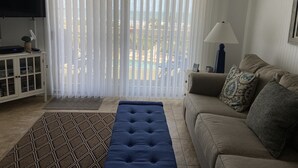 Un salon avec un canapé, un pouf bleu, un téléviseur posé sur un meuble et une vue sur l’extérieur à travers des rideaux transparents.