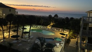 Un espace piscine aménagé sur le toit, avec des chaises longues et des parasols, offrant une vue sur une plage et l’océan au coucher du soleil.
