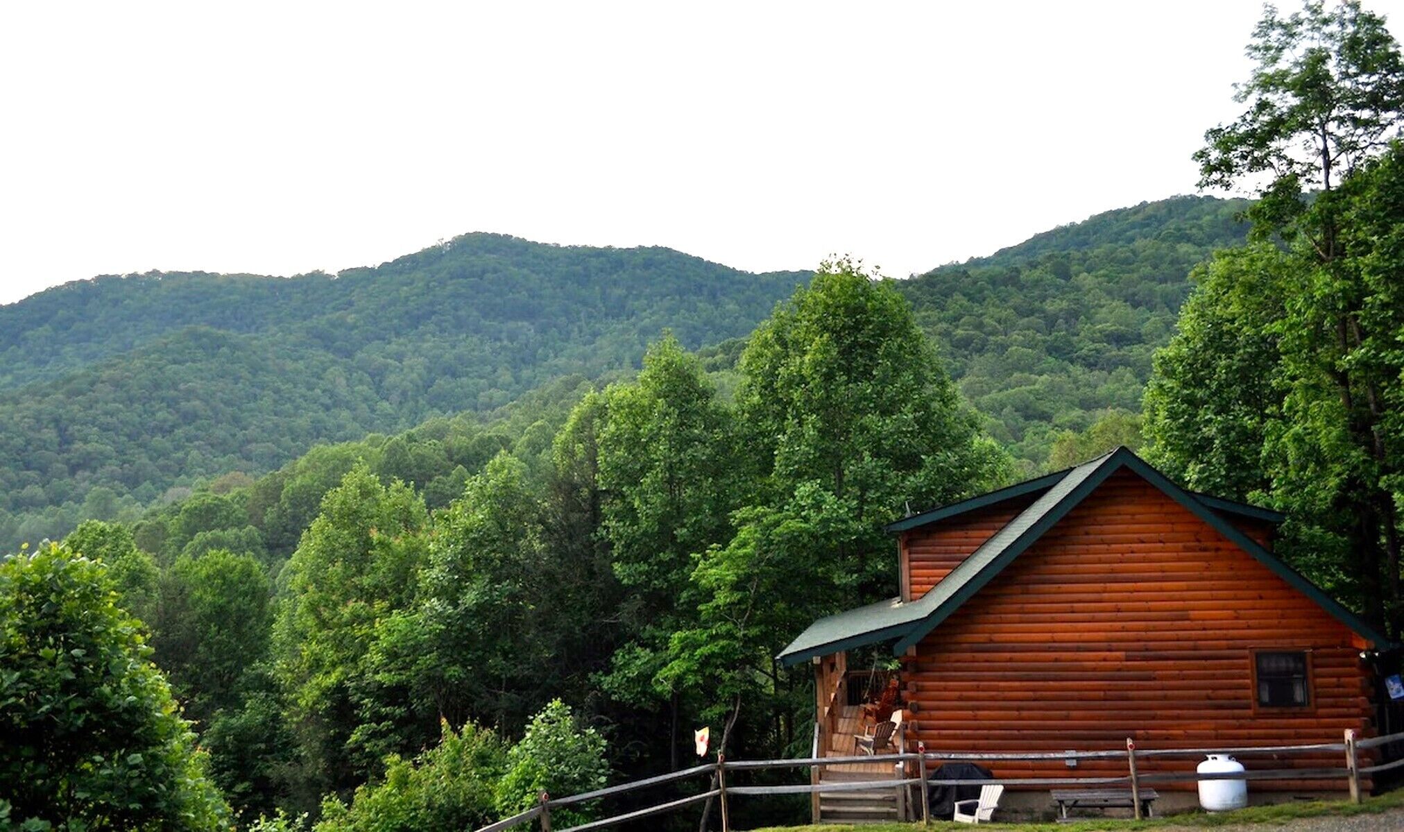 A wooden cabin with a porch, surrounded by lush green forest and mountains.
