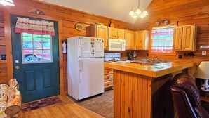A cozy cabin-style kitchen with wooden cabinets, a refrigerator, a microwave, and a dining area with a table and chairs. - Deer Country Vacation Cabins #3 - Long Range View, Dogs Friendly (Waynesville)