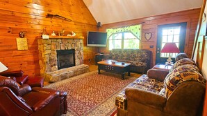 A cozy log cabin interior with a stone fireplace, leather sofas, and a television. - Deer Country Vacation Cabins #3 - Long Range View, Dogs Friendly (Waynesville)