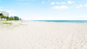 Ein weiter, sandiger Strand mit blauem Himmel und vereinzelten Wolken. Im Hintergrund sind Gebäude und Palmen zu sehen.