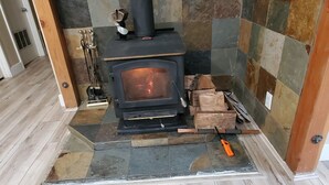A wood-burning stove with a caution sign, a firewood pile, and a tool on the floor.