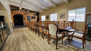A dining area with a long wooden table and chairs, a kitchen area with modern appliances, and large windows providing natural light.