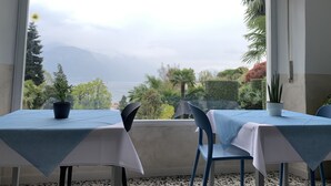 A dining area with two tables, each covered with a blue tablecloth, and chairs. The room has a large window offering a view of a mountainous landscape and a body of water. - Hotel Loveno (Menaggio)