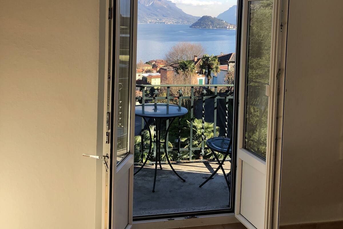 An open sliding door leading to a balcony with a table and chairs, overlooking a lake and mountains.