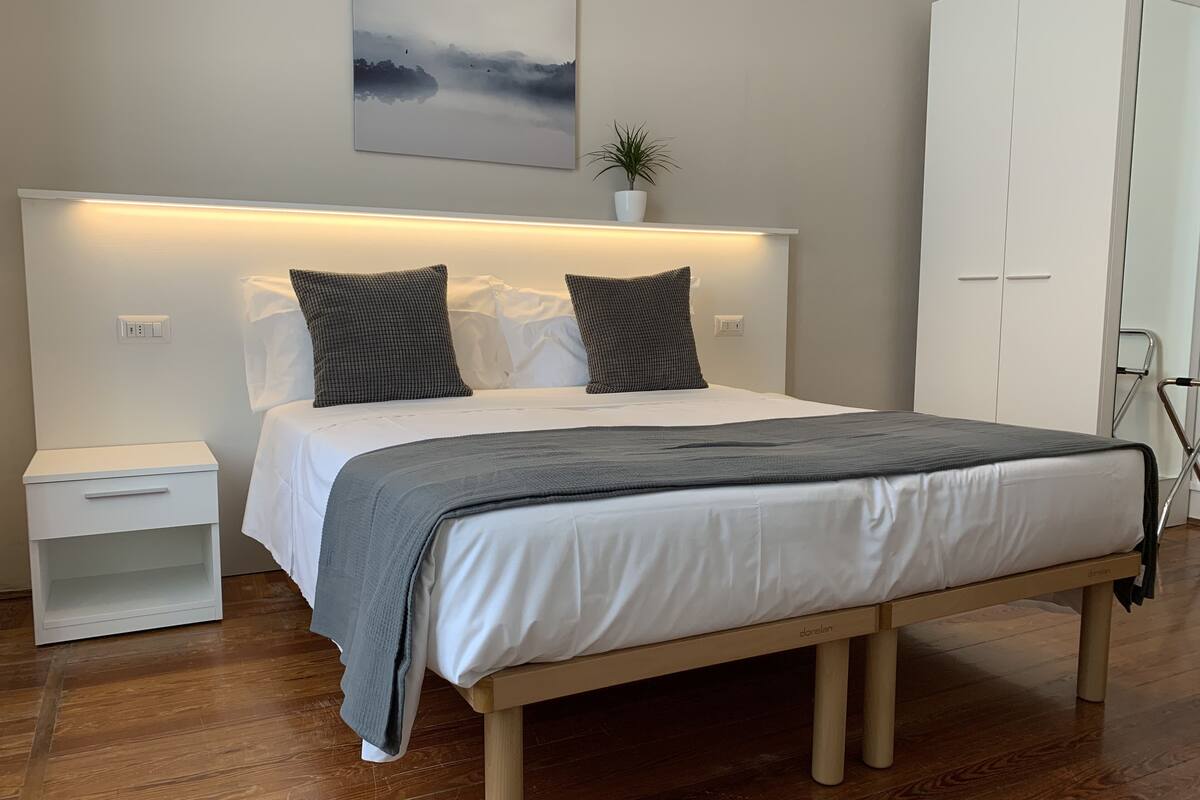 A modern bedroom with a wooden bed, white nightstands, a bedside table, a wall-mounted shelf with a plant, and a wardrobe.