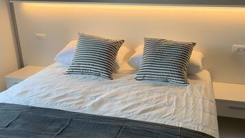 A neatly made bed with white bedding and two patterned pillows, a dark grey throw blanket, and a white nightstand with a power outlet.