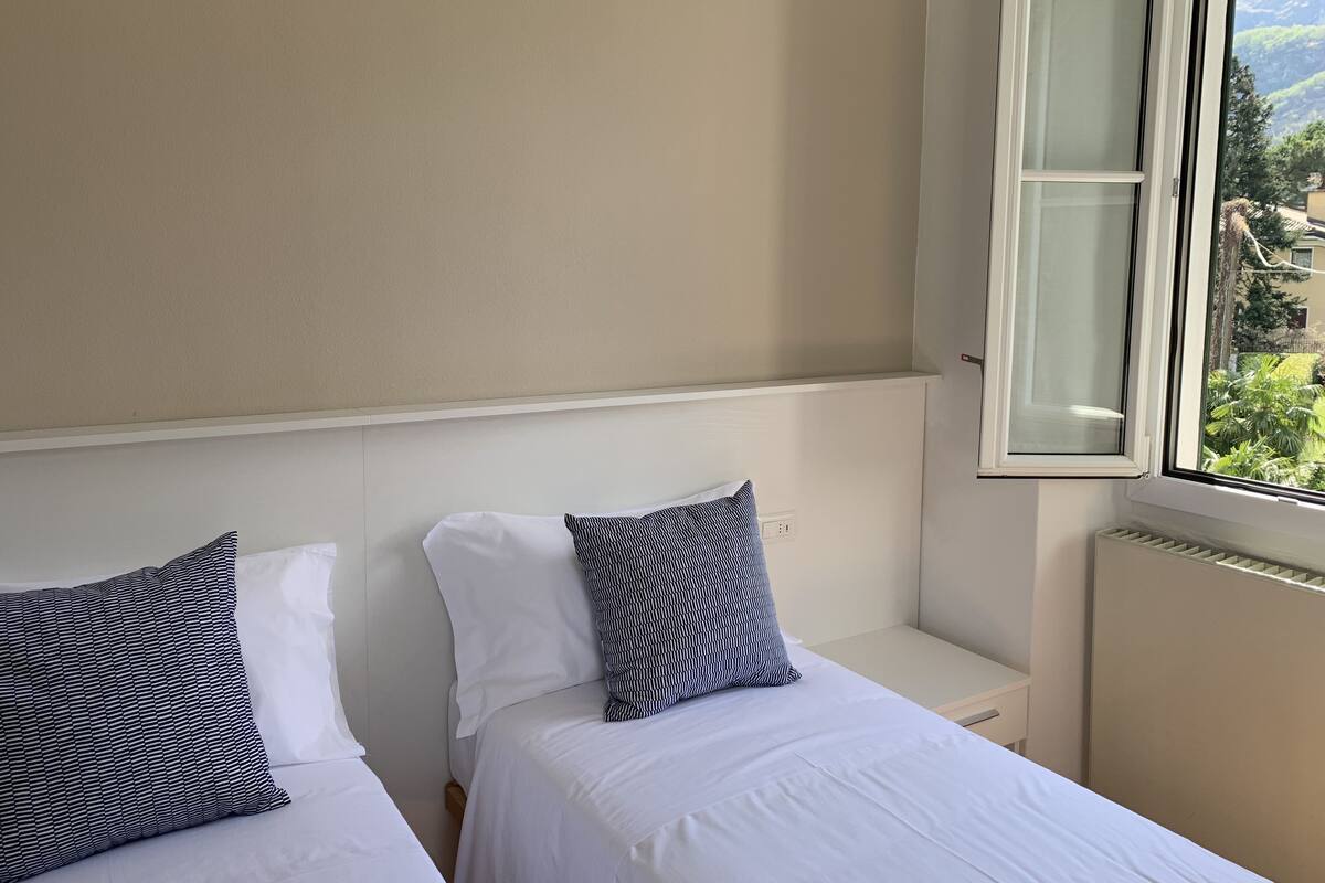 A bedroom with two beds, a window with a view of greenery and mountains, and a white radiator.