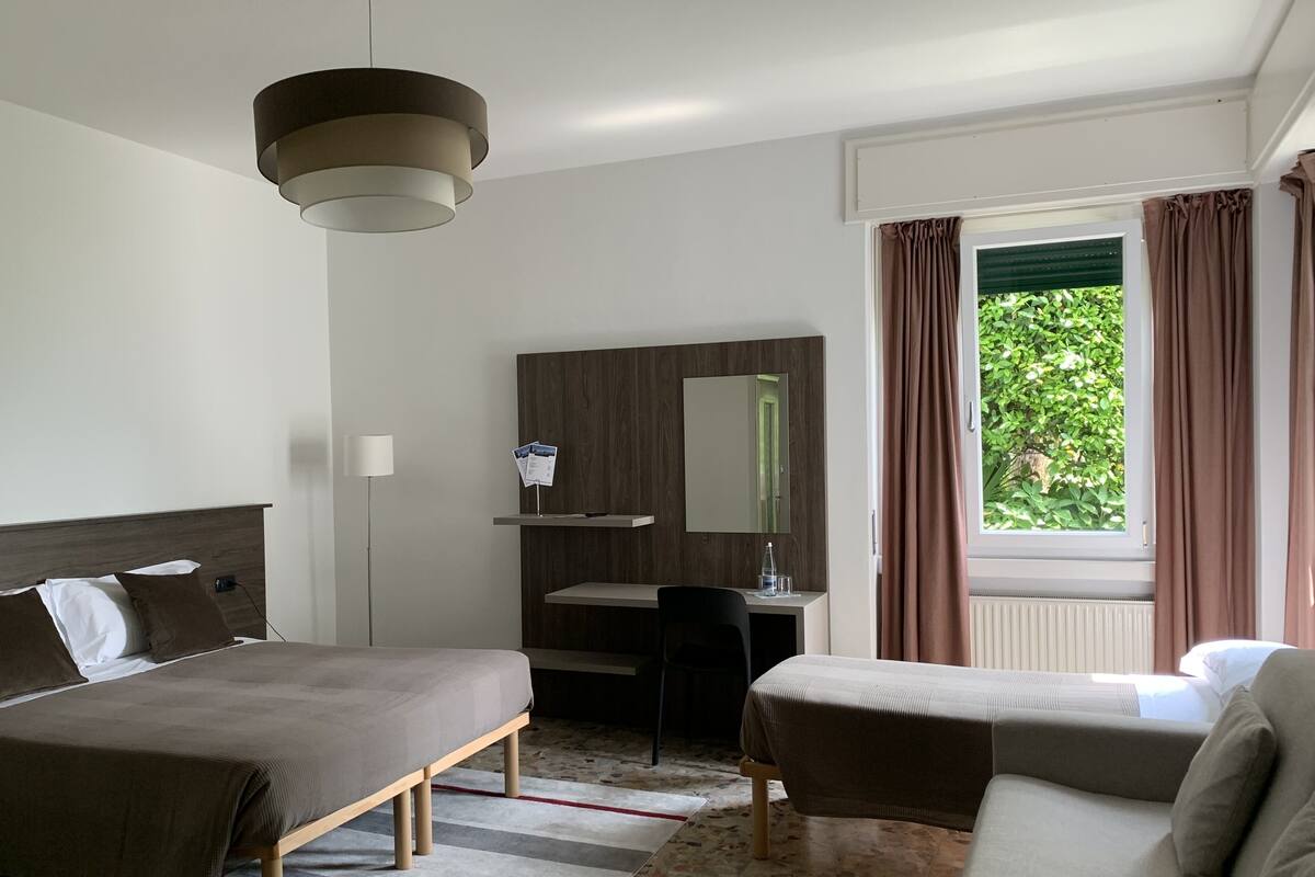 A hotel room with a bed, a sofa, a desk with a chair, a mirror, and a window with curtains.