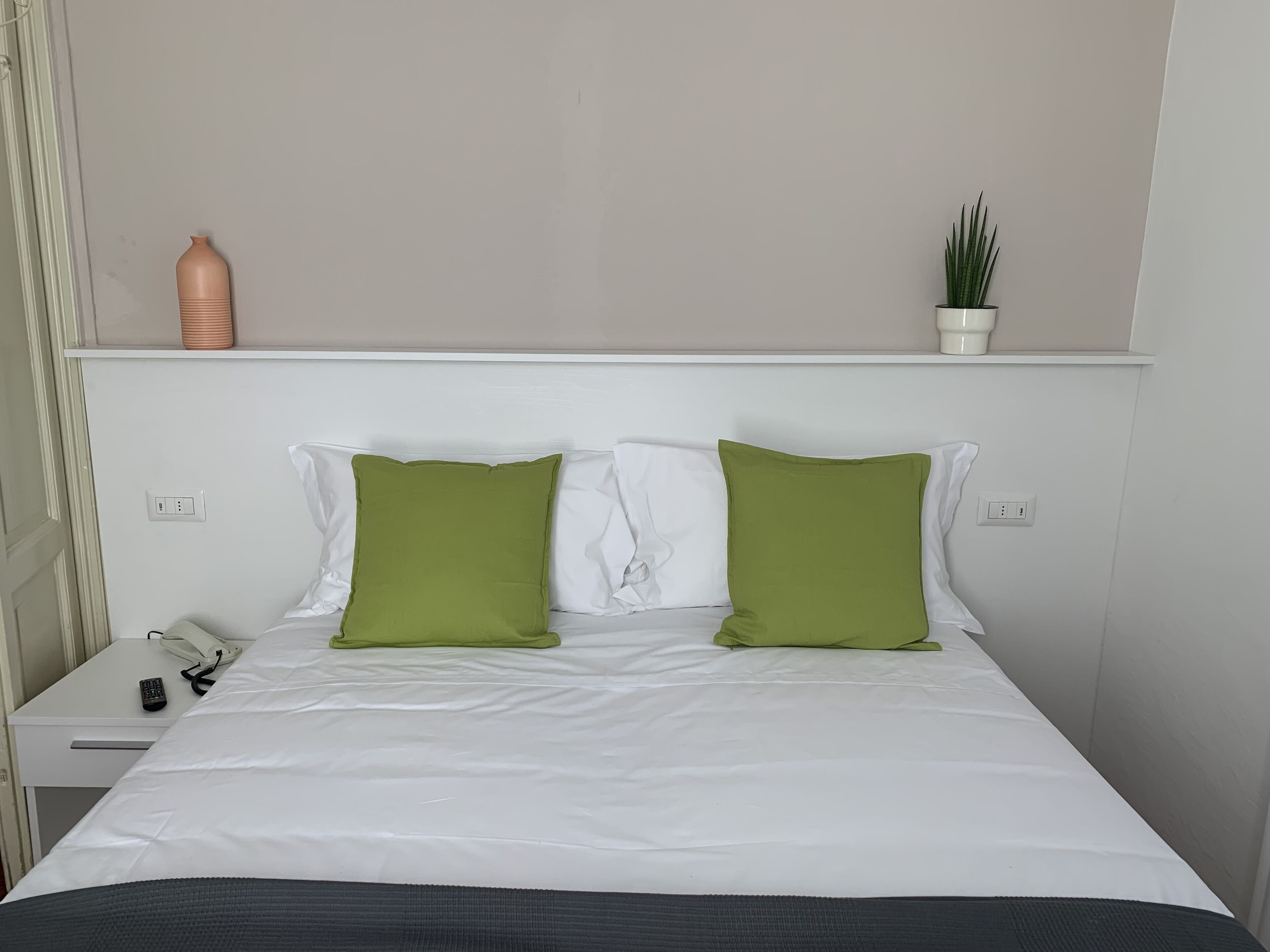 A neatly made bed with white bedding and two green pillows, a bedside table with a phone and remote control, and a small potted plant on the nightstand.
