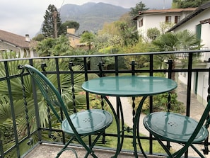 A balcony with two green metal chairs and a glass table, overlooking a garden with various plants and a mountainous backdrop. - Hotel Loveno (Menaggio)