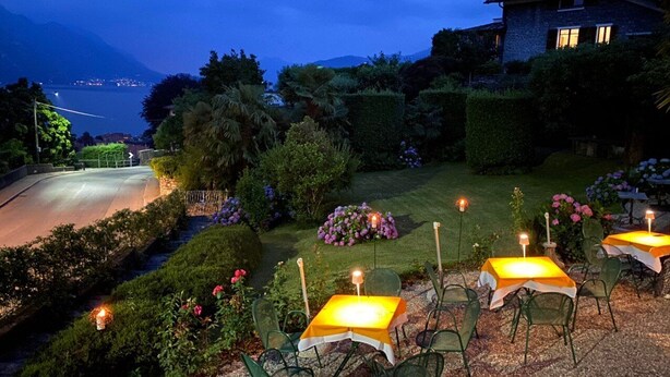 An outdoor dining area with tables and chairs, surrounded by well-maintained gardens and a view of a mountain and lake at dusk. - Hotel Loveno (Menaggio)