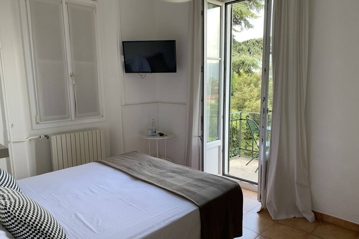 A bedroom with a bed, a television, a radiator, a small table with a bottle, and a balcony with a chair.