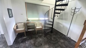 Habitació amb terra de rajoles fosques, una cadira, una taula petita, un mirall i una escala amb barana metàl·lica.
