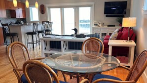 A modern kitchen with wooden cabinets, a dining area with wicker chairs and a glass table, and a living area with a TV and red cushions. - Elegant oceanfront home on Southwest Harbor minutes to trails and village center (Southwest Harbor)