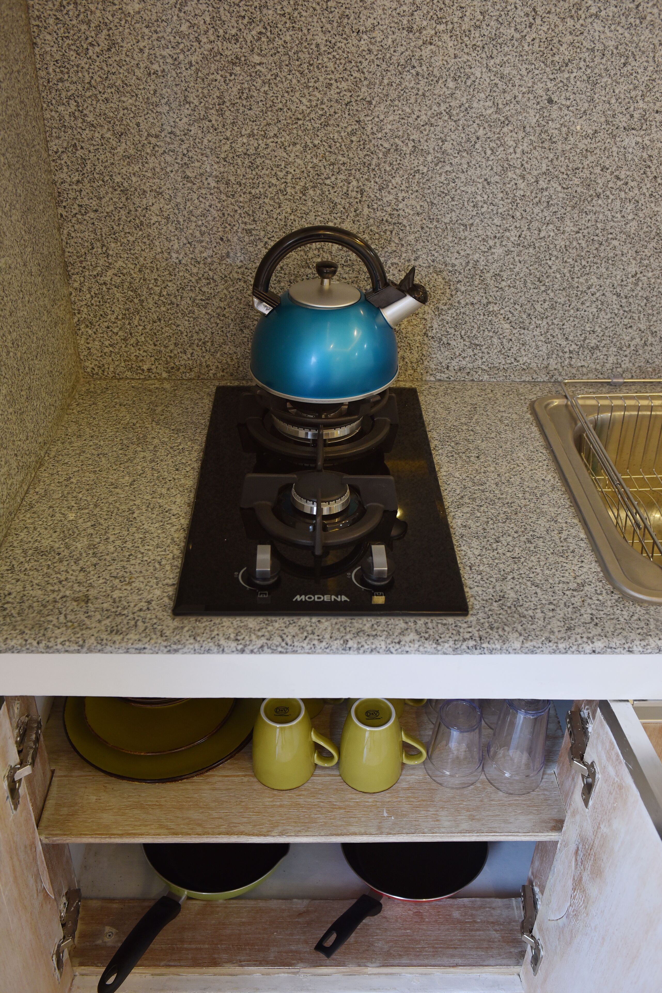 Una cocina con un hervidor azul sobre una estufa negra, un lavamanos y un mueble de madera debajo con platos y tazas.