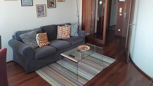 A living room with a grey sofa, patterned cushions, a glass coffee table, and a striped rug. There are framed pictures on the wall and a wooden door in the background. - Porto beach apartment (Matosinhos)