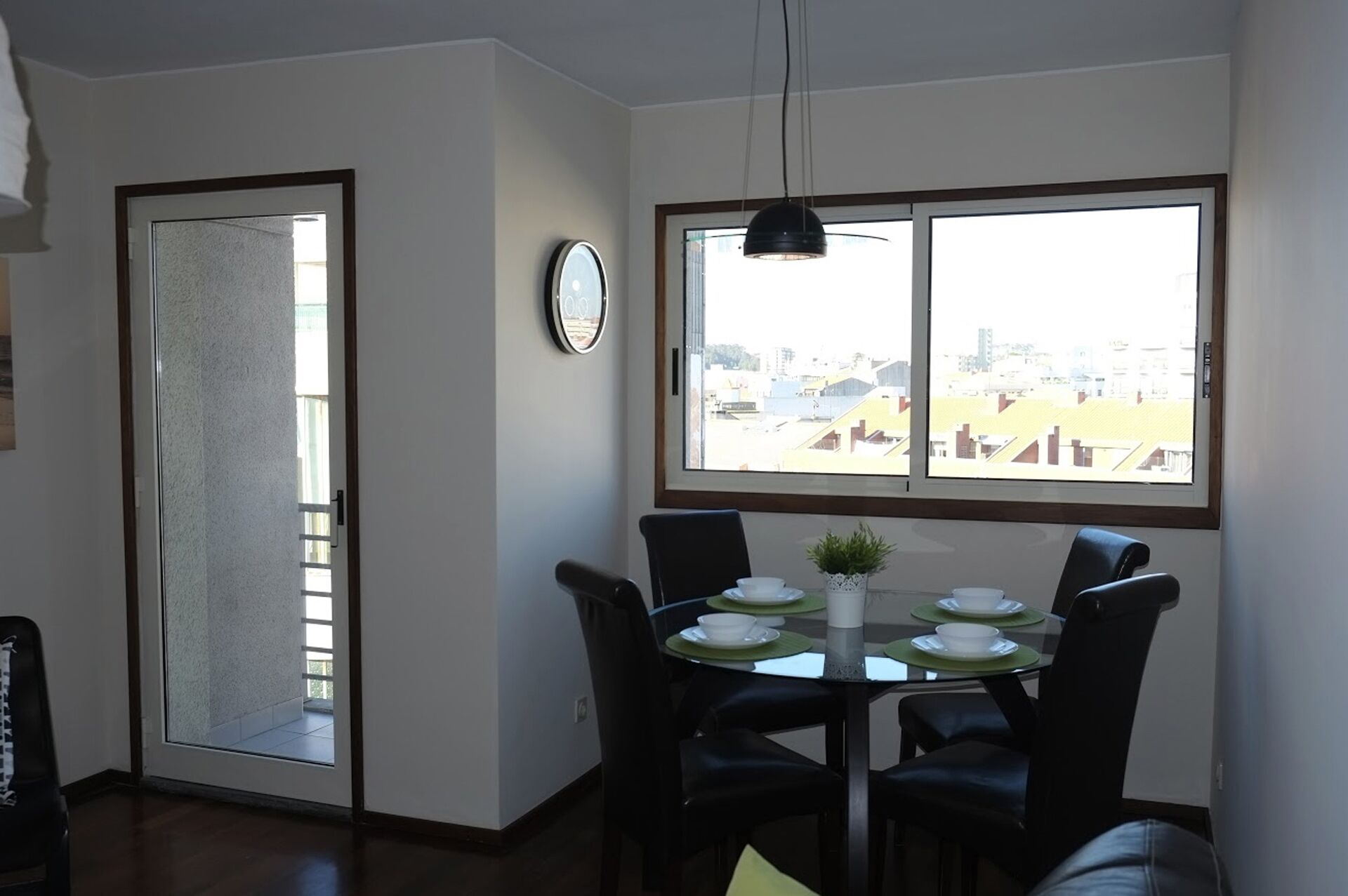 A dining area with a table set for four, a clock on the wall, and a view of buildings through a large window.