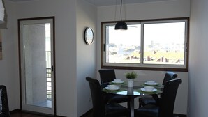 A dining area with a table set for four, a clock on the wall, and a view of buildings through a large window. - Porto beach apartment (Matosinhos)