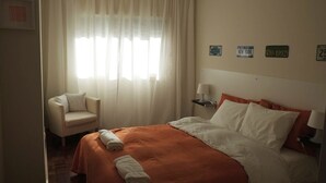 A hotel room with a bed, a chair, and a window with sheer curtains. There are wall decorations and a lamp on a bedside table. - Porto beach apartment (Matosinhos)