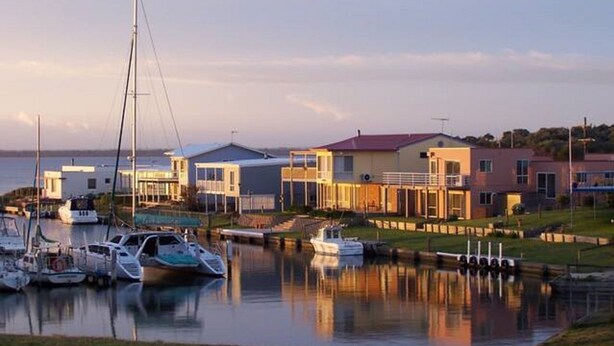Un puerto deportivo con varias embarcaciones amarradas, una casa moderna con techo rojo y una masa de agua tranquila que refleja los edificios.