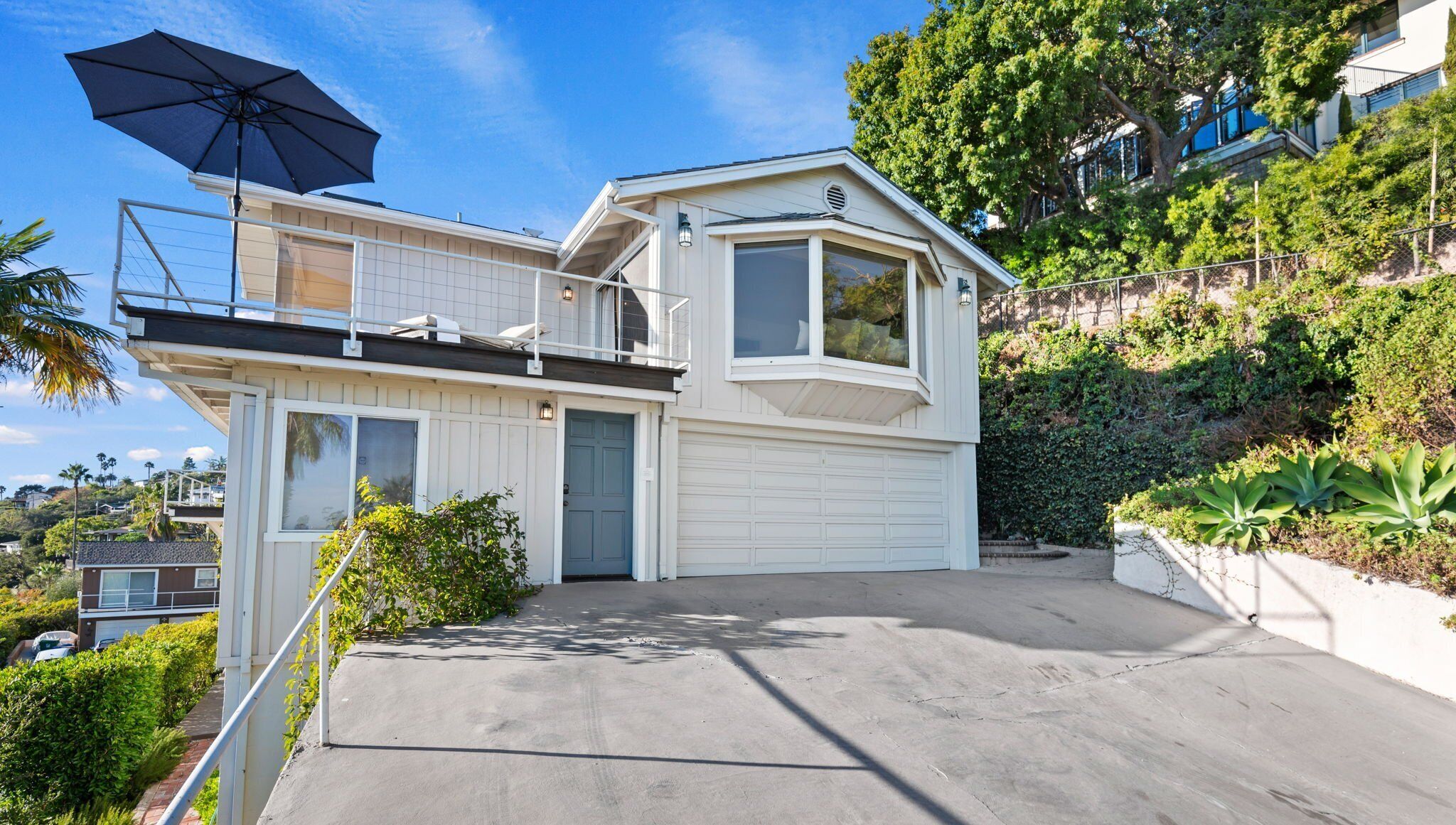 A two-story house with a garage, a balcony with a black umbrella, and a hillside garden.