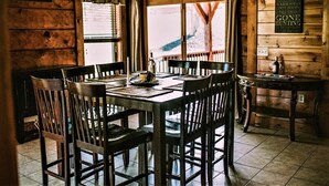 Uma área de jantar rústica em madeira com uma mesa posta para quatro pessoas, um espaço de bar e um aviso indicando as comodidades da pousada.