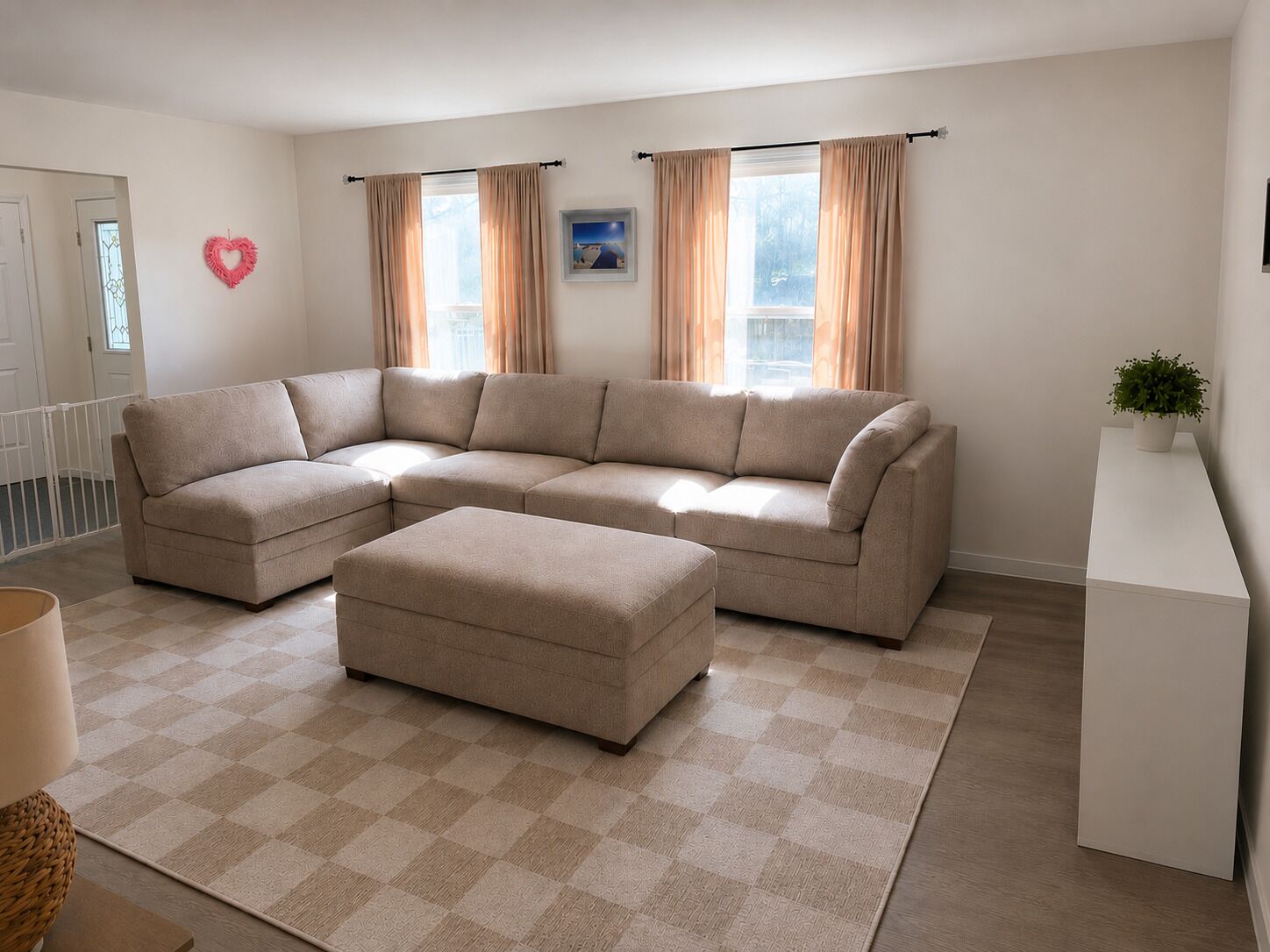 A living room with a beige sectional sofa, a matching ottoman, a checkered rug, a white console table with a plant, and a heart-shaped decoration on the wall.