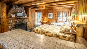 A log cabin room with a stone fireplace, a bed with a quilt, a desk with a lamp, and a window with curtains.