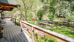 A wooden deck with outdoor seating, a picnic table, and a barbecue grill, overlooking a natural landscape with trees and a stream.
