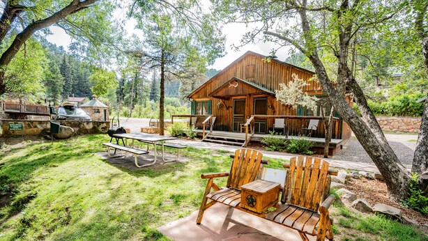 A rustic cabin with a porch, a picnic table, and wooden chairs in a wooded area.