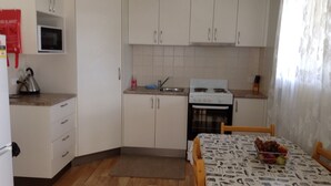 Une cuisine compacte avec des meubles blancs, une cuisinière, un évier et un four à micro-ondes. Un petit tapis est posé sur le sol, et une table est agrémentée d’un panier de fruits.