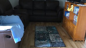 Un salon avec un canapé en cuir marron, un coussin bleu, un meuble en bois rempli de livres et un tapis à motifs posé sur un plancher en bois.