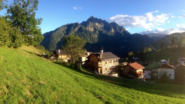 Ein Bergdorf mit traditionellen Holzhäusern, ein markanter Berg im Hintergrund und ein grasbewachsener Hang.