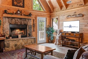 A cozy cabin interior with a stone fireplace, wooden walls, a television, and a teddy bear. - Premium Rustic Cabin Perfect Getaway, Private Hot Tub,Piano, Near Shows & Big Cedar, Jacuzzi Tub! (Ridgedale)