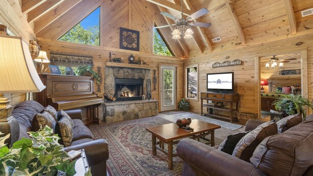 A cozy log cabin interior with a stone fireplace, leather sofas, a television, and a ceiling fan. - Premium Rustic Cabin Perfect Getaway, Private Hot Tub,Piano, Near Shows & Big Cedar, Jacuzzi Tub! (Ridgedale)