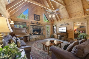 L’intérieur d’un chalet confortable, avec une cheminée en pierre, des canapés en cuir, un téléviseur et un ventilateur de plafond.