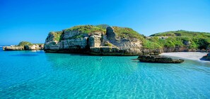 A coastal scene with clear turquoise waters, rocky formations, and a sandy beach.