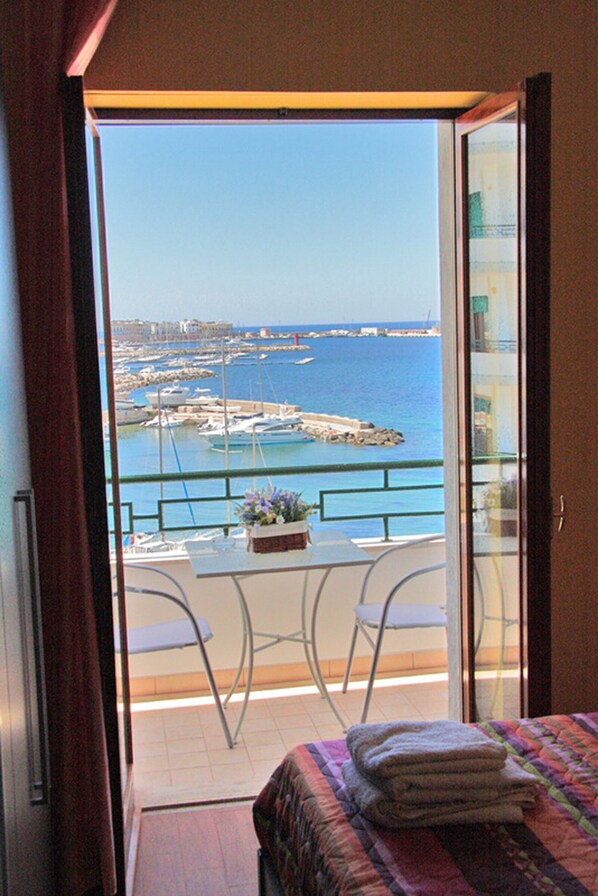 A balcony with a view of the sea and boats, a small table with chairs, and a bed with a folded blanket.