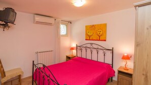 A bedroom with a metal bed frame, a red bedspread, two bedside lamps, an air conditioner, a television mounted on the wall, and a window with a curtain. - Borghetto Tio Pepe - Trilo 6 Mandarin (Mattinata)