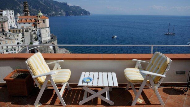 A balcony with two striped chairs and a small table, overlooking a coastal town with historic buildings and the sea. - ATRANI "CASA ROSSA" AMALFI COAST WITH SEA VIEW (ATRANI)